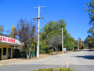 Shows the view looking up Pack Road in Marysville in Victoria. In the left of the photograph can be seen a Ski Hire shop and further up the road is a wooden sign.
