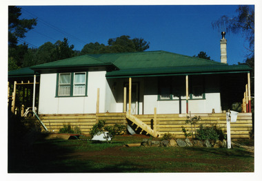 Shows a cream and green house. A verandah can be seen around three sides with a garden bed at the front. There is also a letter box with the number 108 attached.