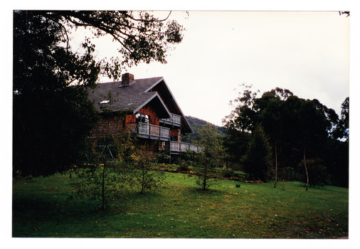 Shows a timber clad double storey house set in a garden of lawn and trees.