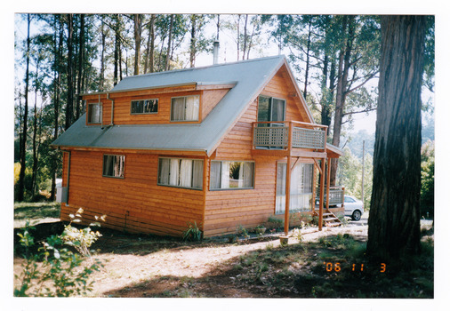 Shows a wooden A-frame house surrounded by trees. There is a white car parked out the front of the house.