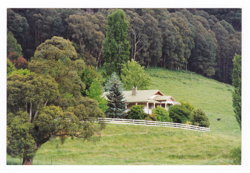 Shows a house surrounded by trees and a wooden fence. In the background is a heavily forested hill.