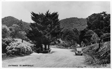 Shows the view looking up the Marysville-Wood's Point Road towards the corner with Murchison Street in Marysville in Victoria. Shows an unsealed road leading off to the left. There is a glimpse of another road leading off to the right. In the foreground there are shrubs and large trees on the left hand side of the road and on the right there is a parked car. In the background are heavily forested mountains. The title of the photograph is handwritten in black ink on the lower edge.