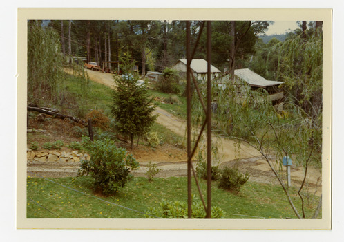 Shows an intersection of two dirt roads. One road leads past two buildings, one of which is a double storey house with verandahs out the front. In the foreground of the photograph is a blue letter box.