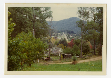 Shows the view from a high location looking towards a heavily forested mountain. In the foreground is a dirt track leading past a house and a small wooden shack. In the distance is a road leading into the distance with buildings on either side.