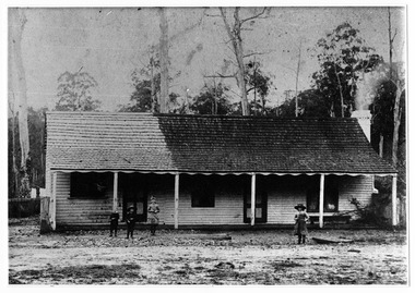 Shows a weatherboard building with a timber shingled roof. Across the front of the building is a verandah which has a bench seat set on it. The building also has a brick chimney. In front of the building are four children.