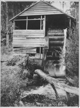 Shows a wooden building which houses a water wheel.