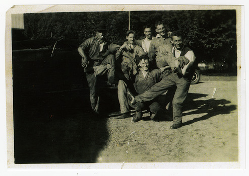 Shows a group of six men standing and kneeling in front of a truck. In the background can be seen the front right hand wheel of another vehicle.