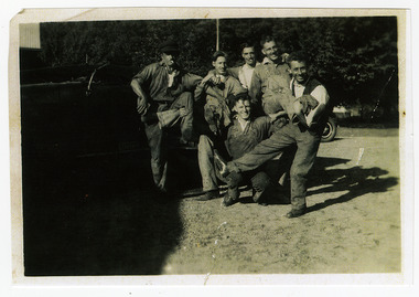 Shows a group of six men standing and kneeling in front of a truck. In the background can be seen the front right hand wheel of another vehicle.