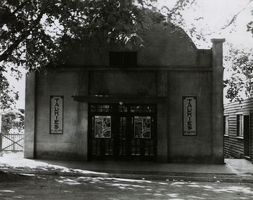 Shows a black and white photograph of the Marysville Theatre in Marysville in Victoria. Shows a large building constructed of concrete. At the centre of the front of the building are large framed glass paneled doors. On the front facade at either side of the doors are two signs advertising 'Talkies'. The roof of the building has a curved gable at the front. 