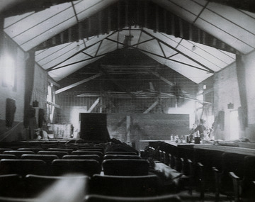 Shows the inside of the Marysville Theatre in Marysville in Victoria. Shows the building in the process of being converted from a picture theatre. In the background can be seen a man sitting on a frame of scaffolding who appears to be talking to another man who is standing in the right of the photograph. The rows of chairs for the theatre patrons can be seen.