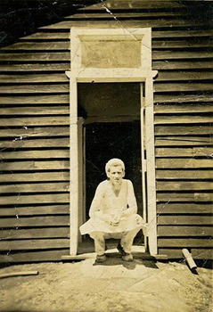 Show a man wearing an apron and a cap crouched in a doorway of a wooden building.