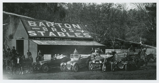 Shows a large barn like building with Barton Bros Stable painted on the roof in large letters. In front of the building are four early model cars, all with people in them. One of the cars has three ladies seated in the rear passenger seat. There are also three riding horses as well as three horse drawn carts and carriages; all have riders, drivers and passengers.