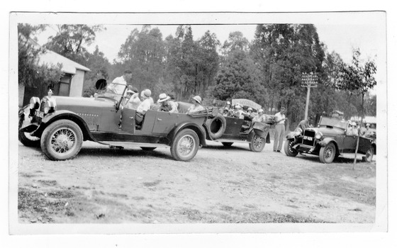 Shows three early model cars all carrying several passengers. All the cars are parked outside an unknown building. Behind the last car is an electricity pole.