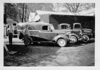 Shows three small trucks parked outside a building. Standing next to one of the trucks are two young men. In the background can be seen another vehicle.