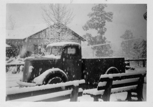 Shows a small truck parked next to a building. It is snowing quite heavily and the truck and its surrounds are covered in snow.