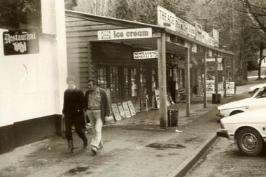 Shows the street front view of Barton Bros' Store in Marysville in Victoria with the footpath running along the front. There are two men walking along the footpath with cars parked in front of the store. Against the window of the shop are several newpaper headlines. In the background are more buildings with signage in front of them.