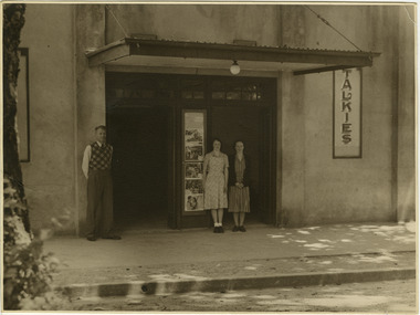 Shows a black and white photograph of Jim, Marj and Marie Wallace standing in front of the Marysville Theatre in Marysville in Victoria.