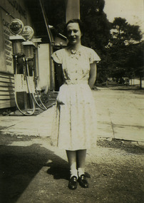 Shows Jill Wallace standing outside the McKenzies Bus Tourist Depot in Marysville in Victoria. In the background are the petrol pumps for the depot.