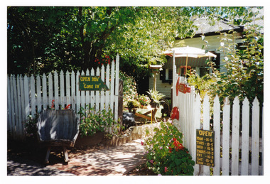 Shows the cream picket fence that was outside Past Favorites in Marysville in Victoria. Attached to the fence are an open sign and the opening hours of the business. A view of the house can be seen beyond the fence.
