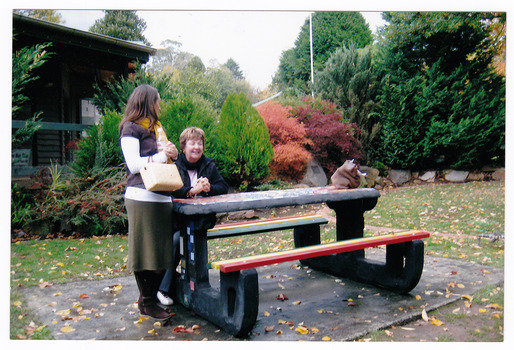 Shows a decorated wooden table with bench seats outside the Marysville Visitor Information Centre in Victoria. Shows one lady sitting at the table talking to another lady who is standing next to the table.