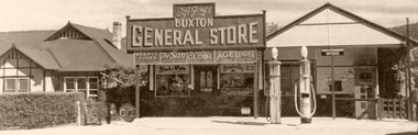 Shows the view from across the road towards a small house beside the Buxton General Store. Shows a weatherboard building with parapet at roof with lettering: Buxton General Store and signs for newspapers on the windows. Adjacent to the store is the Buxton Post Office that has two petrol bowsers at the front of the building.