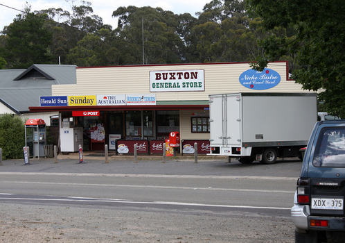Shows the Buxton General Store in Victoria. Above the front of the store is a sign with the name of the store as well as a sign advertising The Niche Bistro and Court Yard. There is a red Post Office box out the front. There is also a telephone paybox outside the store. Shows a delivery truck out the front of the store.