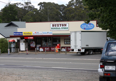 Shows the Buxton General Store in Victoria. Above the front of the store is a sign with the name of the store as well as a sign advertising The Niche Bistro and Court Yard. There is a red Post Office box out the front. There is also a telephone paybox outside the store. Shows a delivery truck out the front of the store.