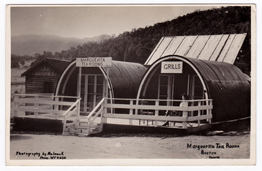 Shows the buildings of the Marguerita Tea Rooms in Buxton in Victoria. Shows two curved roofed buildings with a verandah running along their fronts. At the left of those buildings is a wooden building and behind them is a large timber roofed building. On both of the curved buildings are advertising signs for the Marguerita Tea Rooms and Grills. In front of one of the buildings there is a lady sitting on a chair on the verandah.