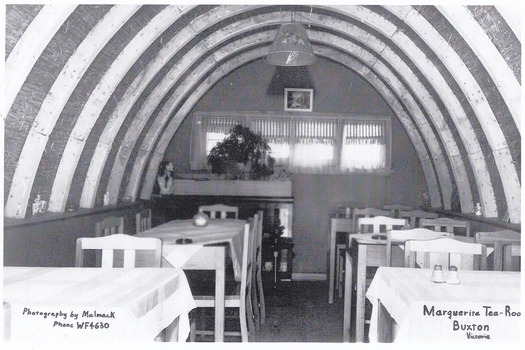 Shows the interior of the Marguerita Tea Rooms in Buxton in Victoria. Shows the tables and chairs with table cloths on some of the tables. Shows the curved roof line of the building with a long narrow window at the rear of the room.