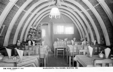 Shows the interior of the Marguerita Tea Rooms in Buxton in Victoria. Shows the tables and chairs with checked table cloths on the tables. There are also glasses with table napkins folded inside the glasses. Shows the curved roof with a long narrow window at the rear of the building.