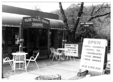 Shows the front facade of the Olde Yarra Track Shoppe in Marysville in Victoria. Shows three advertising signs advising what goods are available. Out the front are two table with chairs. There is a sign above the doorway with the name of the shop. 