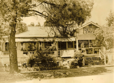 Shows the front facade of the General Store and Cafe in Marysville in Victoria. Shows a large weatherboard building with stairs leading up to a small verandah. The roof is of corrugated iron. The front windows all have shutters attached. There are several advertising signs adorning the front windows of the building. There is a sign advertising the Pine Tree Tearooms and what foods and services it offers.