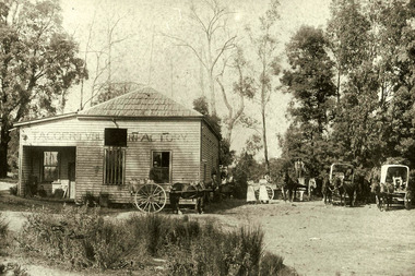 Shows the Taggerty Butter Factory in Victoria. Shows a weatherboard building with wooden shingles on the roof. The name of the factory is along the front of the building. Standing in front of the building is a horse driven cart with several milk cans on board as well as the carter. Standing next to the building are two butter factory workers who are both wearing caps and long aprons. There are also four horse drawn delivery wagons waiting next to the building, two of which have canopies over the wagons.