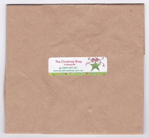 Shows a brown paper bag with a sticker adhered to it. The sticker shows details of The Christmas Shop in Marysville; a mobile telephone number and the website address.