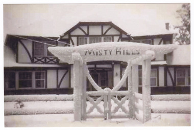 Shows a tudor style building behind a fence with an ornate gateway. The name of the guest house is along the top of the gateway. The guest house, fence and gateway are all covered with a heavy covering of snow, as is the surrounding ground.
