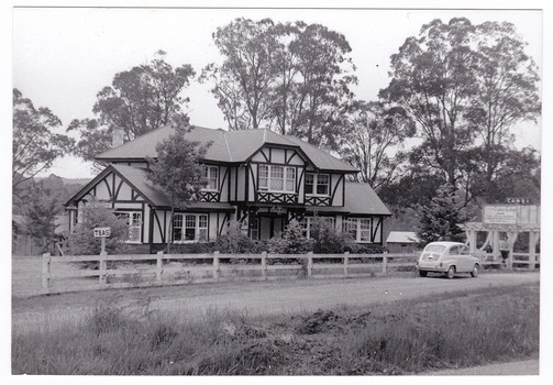 Shows the Tudor Lodge Cafe in Narbethong in Victoria. Shows a large tudor style building that has a fence along the front with an ornate wooden gateway. There is a car parked in front of the gate. There is a large advertising sign attached to the gateway.