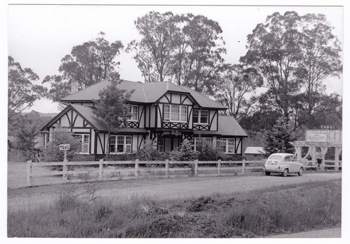 Shows the Tudor Lodge Cafe in Narbethong in Victoria. Shows a large tudor style building that has a fence along the front with an ornate wooden gateway. There is a car parked in front of the gate. There is a large advertising sign attached to the gateway.