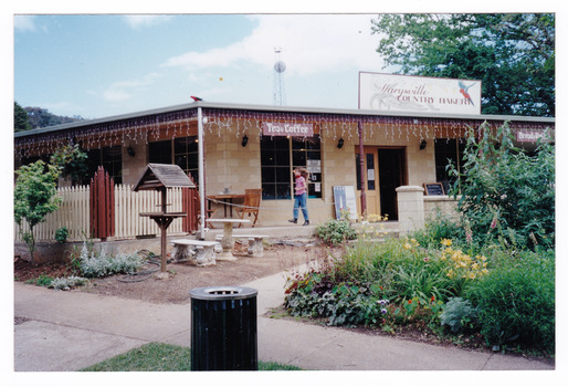 Shows the front facade of the Marysville Country Bakery in Victoria. Shows the name of the bakery on a large sign that sits above the verandah. Shows a set of table and chairs out the front.
