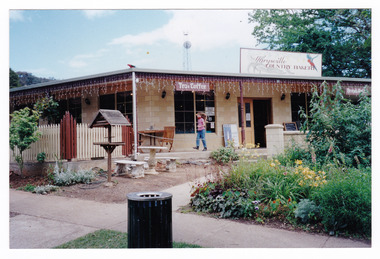Shows the front facade of the Marysville Country Bakery in Victoria. Shows the name of the bakery on a large sign that sits above the verandah. Shows a set of table and chairs out the front.