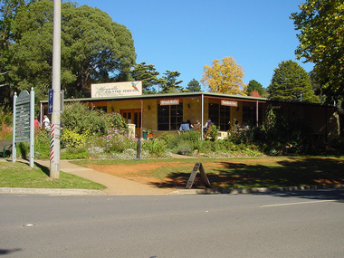 Shows the front facade of the Marysville Country Bakery in Victoria. Shows a large sign with the name of the bakery above the verandah. Hanging under the verandah are signs advertising bread rolls and sandwiches. There is a group of people sitting at a table and chairs on the verandah.
