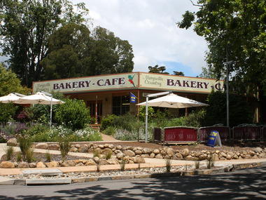 Shows the front facade of the Marysville Country Bakery in Victoria. Shows on large signs above the verandah the name of the bakery. Out the front of the building are gardens with shade umbrellas above seating.