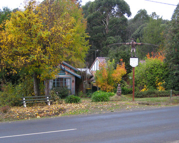 Shows the front facade of Bruno's Art and Sculptures Garden in Marysville in Victoria. Shows two weatherboard buildings with a signpost out the front. At the top of the signpost is a sculpture of a man who has large wings attached to his arms. He is also wearing a top hat that has a bird sitting on top of it. Near the base of the signpost is a sculpture of a kangaroo with a frog in its pouch.