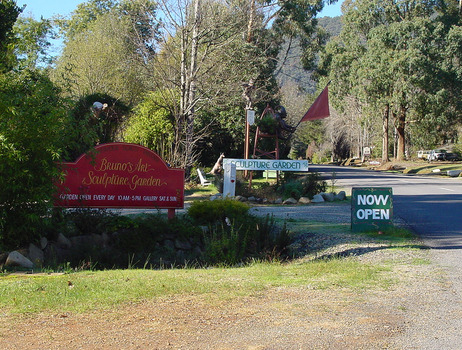 Shows the signs out the front of Bruno's Art and Sculptures Garden in Marysville in Victoria. In the background can be seen several sculptures including one of a kangaroo.