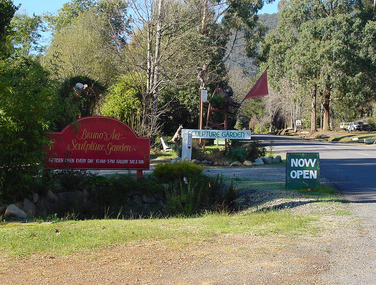 Shows the signs out the front of Bruno's Art and Sculptures Garden in Marysville in Victoria. In the background can be seen several sculptures including one of a kangaroo.