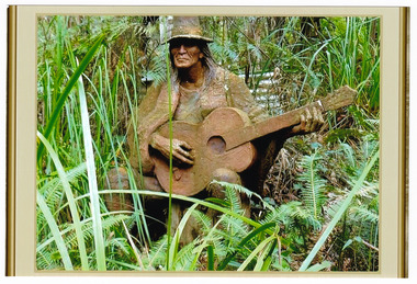 Shows a terracotta sculpture of a man playing a guitar. The man has long hair and is wearing a hat and is wearing a cloak. The man is sitting.