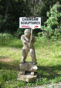 Shows a chainsaw sculpture of a man standing on a section of timber log weilding an axe. Attached to the sculpture is a sign directing visitors to the chainsaw sculpture garden.
