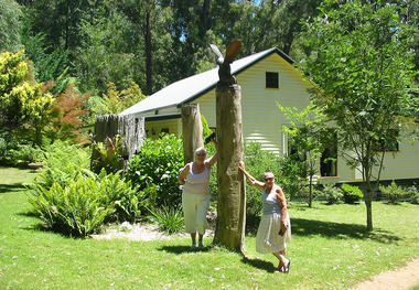 Shows a chainsaw sculptured eagle atop a large wooden pole. Shows two ladies standing either side of the pole with a weatherboard house in the background.