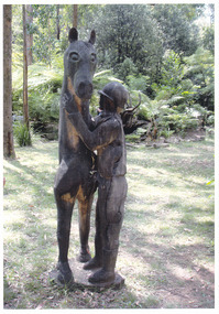 Shows a chainsaw sculpture of a horse with its rider standing next to it. 