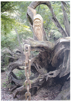 Shows a chainsaw sculpture of a man's face positioned behind a fallen tree. The man has a long beard. Hanging from one of the tree's roots is a wooden linked chain.