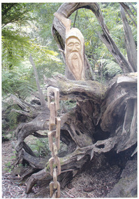 Shows a chainsaw sculpture of a man's face positioned behind a fallen tree. The man has a long beard. Hanging from one of the tree's roots is a wooden linked chain.